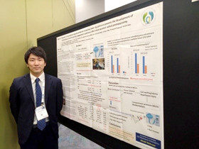 スーツ姿の男性が、大きなポスターの前に立っている様子。