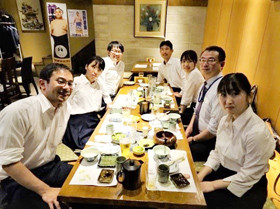 室内のテーブルで白いシャツを着た男女が集まっている様子。