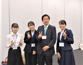 4人のスーツ姿の人々がカメラに向かってピースサインをし、笑顔を見せている様子。