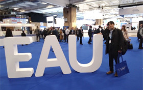 青いカーペットの敷かれた会場で、大きな「EAU」の文字の横に立っている男性。