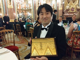 華やかな会場で、タキシード姿の男性が金色の賞状を持っている様子。