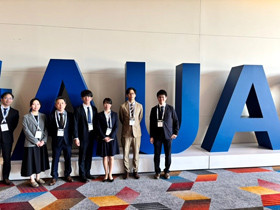 7人のが、巨大な文字オブジェの前でフォーマルな服装で並んで立っている様子。