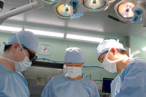 手術室で手術に集中する3人の医師たち。明るい照明と医療機器に囲まれている。