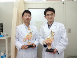 白衣を着た2人の男性が模型を手に持っている様子。