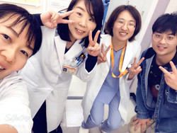 白衣を着た医療従事者とカジュアルな服装の人がピースサインをしながら笑顔で集合写真を撮っている。