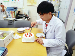 白衣を着た人物が、机の上でケーキを切り分けているシーン。