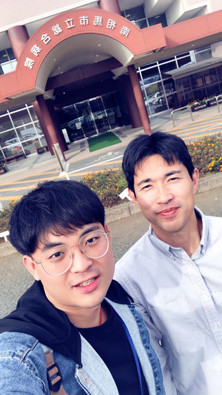 二人の男性が病院の前で笑顔で立っている様子。