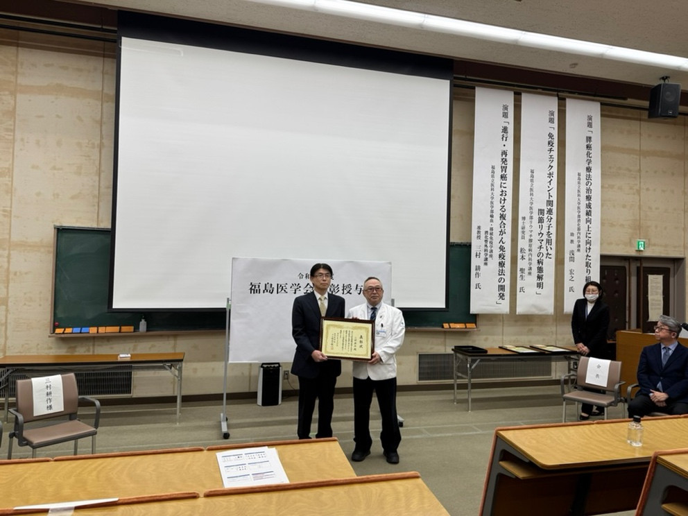 三村耕作准教授が福島医学賞を受賞し、表彰状を手に持っているシーン。背景には講演会の会場が映っている。