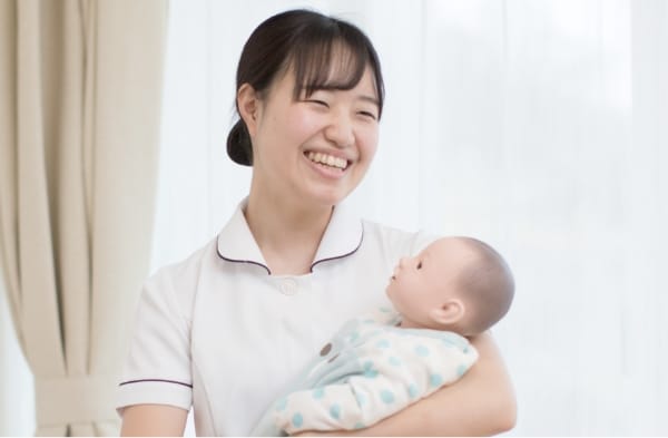 助産師の女性が人形の赤ちゃんを抱き、笑顔でカメラを見つめている様子。背景には明るい窓があり、温かい雰囲気が漂う。