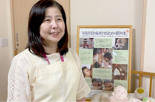 助産師の草野祐香利さんが、助産院の前で微笑みながら立っている様子。背景には助産に関する情報が掲示されたポスターが見える。