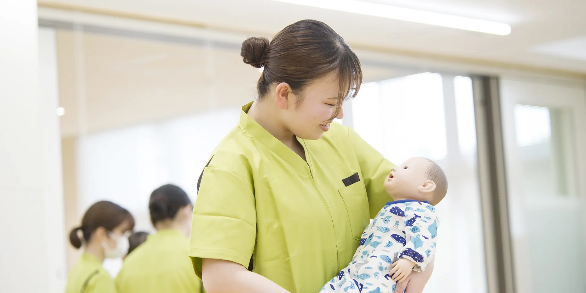助産学専攻の学生が人形の赤ちゃんを抱きしめて微笑んでいる様子。背景には他の学生が見える。