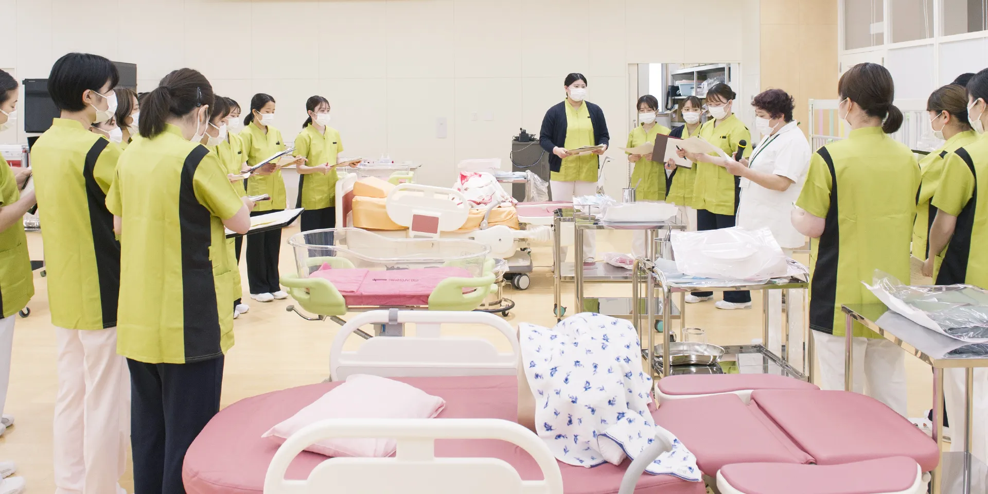 助産学専攻の学生たちが実習室で指導を受けている様子。医療用ベッドや器具が整然と配置されている。