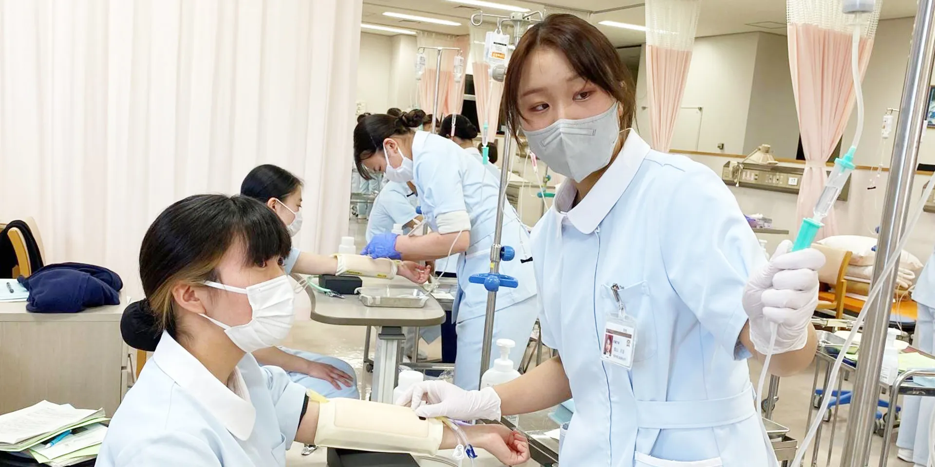 看護学部の実習風景で、学生が患者に点滴を行う準備をしている様子。
