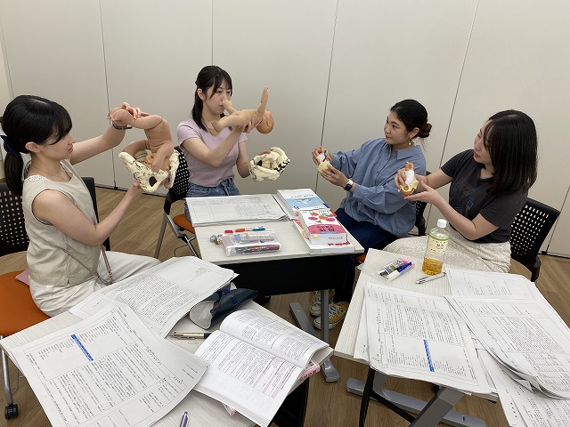 助産学の授業で模型を使いながら学ぶ学生たちの様子。テーブルには資料や文房具が散らばっている。