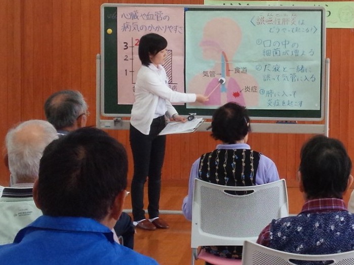 高齢者向けの健康講座で、講師がスライドを指し示しながら説明している様子。受講者は数名、真剣に聞いている。