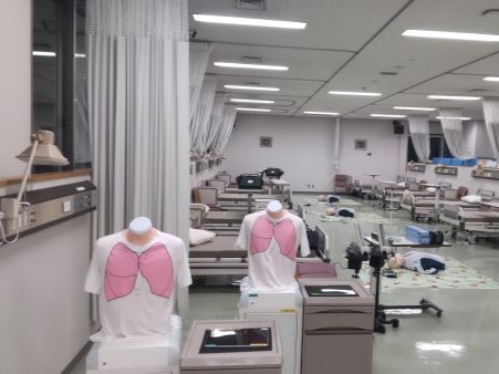 成人看護学の実習室で、胸部のイラストが描かれた白いTシャツを着たマネキンが並ぶ様子。背景にはベッドや医療機器が配置されている。