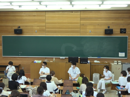 精神看護学の講義中、教員が学生に向けて説明している様子。教室内には多くの学生が座っている。
