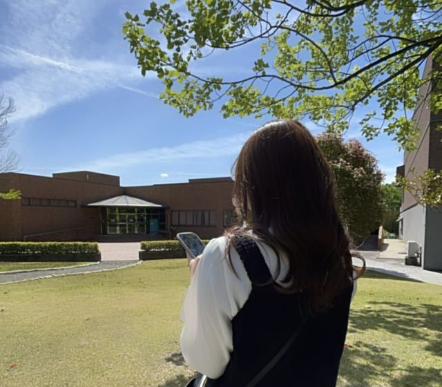 福島県立医科大学のキャンパスでスマートフォンを操作する女性の後ろ姿。青空と緑の木々が広がる風景。