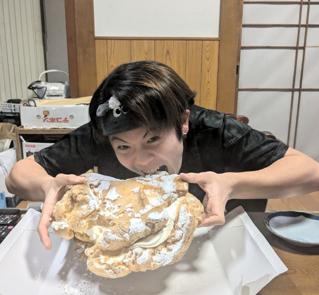 頭に黒いバンダナを巻いた学生が、大きなシュークリームにかぶりつこうとしている姿。