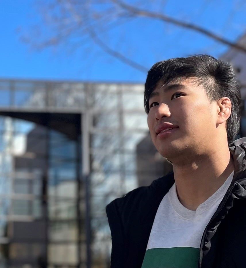 若い男性が青空の下で微笑みながら、近くの建物を背景に立っている様子。