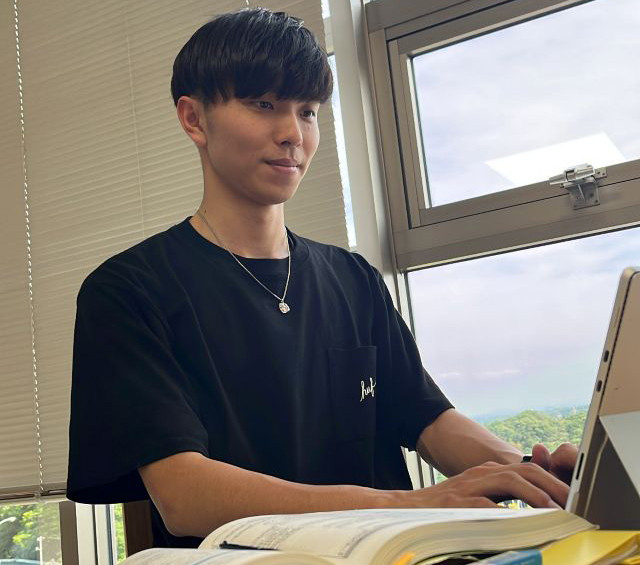 大学の教室で、ノートパソコンを使用して勉強する学生の姿。開いた教科書とノートが机に置かれている。