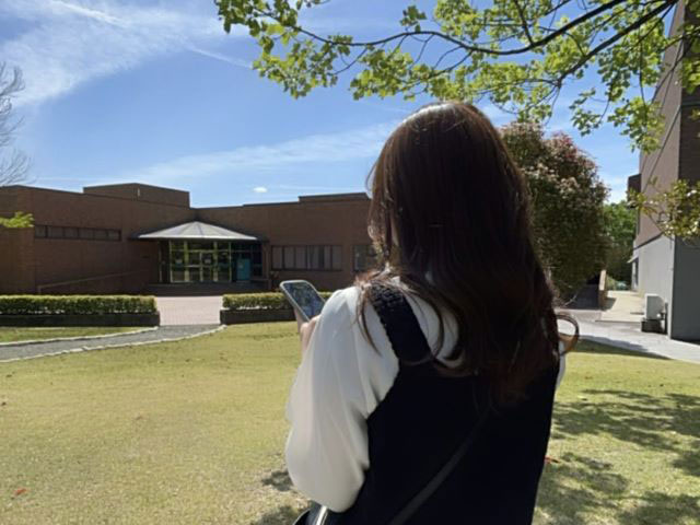 福島県立医科大学のキャンパスで、スマートフォンを持ちながら建物を見つめる女性の後ろ姿。青空と緑が広がる穏やかな日。