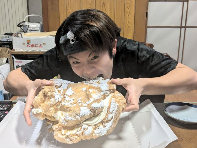頭に黒いバンダナを巻いた学生が、大きなシュークリームにかぶりつこうとしている姿。