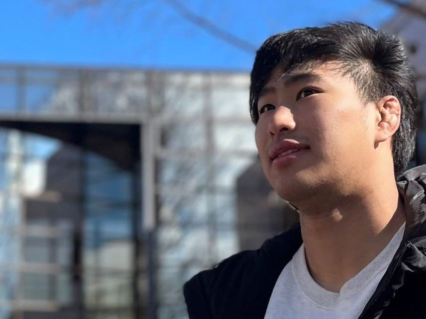 福島県立医科大学のキャンパスで、若い男性が晴れた空の下で考え込んでいる様子。