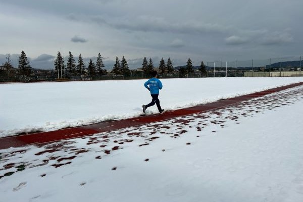 雪に覆われた陸上トラックを走る選手の姿。背景には木々と曇り空が広がる。