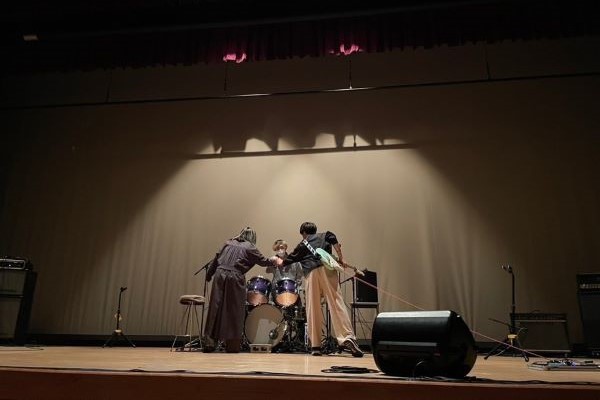 軽音部のメンバーがライブの準備をしている様子。ドラムセットを囲み、照明が当たるステージ上の緊張感が伝わる。