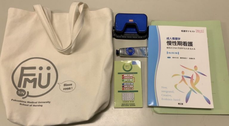 福島県立医科大学のロゴ入りトートバッグ、青いケース、ハンドクリーム、教科書、メモ用紙が並んでいる画像。