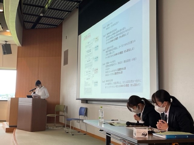 2023年度統合実習報告会でプレゼンテーションを行う学生と、資料を確認する2人の学生の様子。