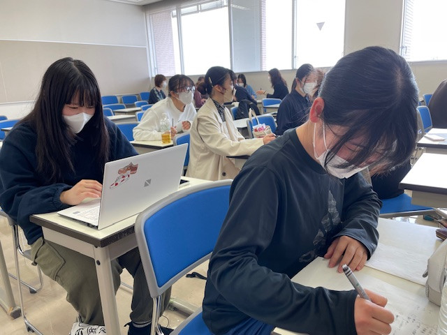 看護学部の教室で、マスクを着用した学生たちが授業に集中している様子。手前の学生はノートにメモを取り、後ろにはパソコンを使っている学生も見える。