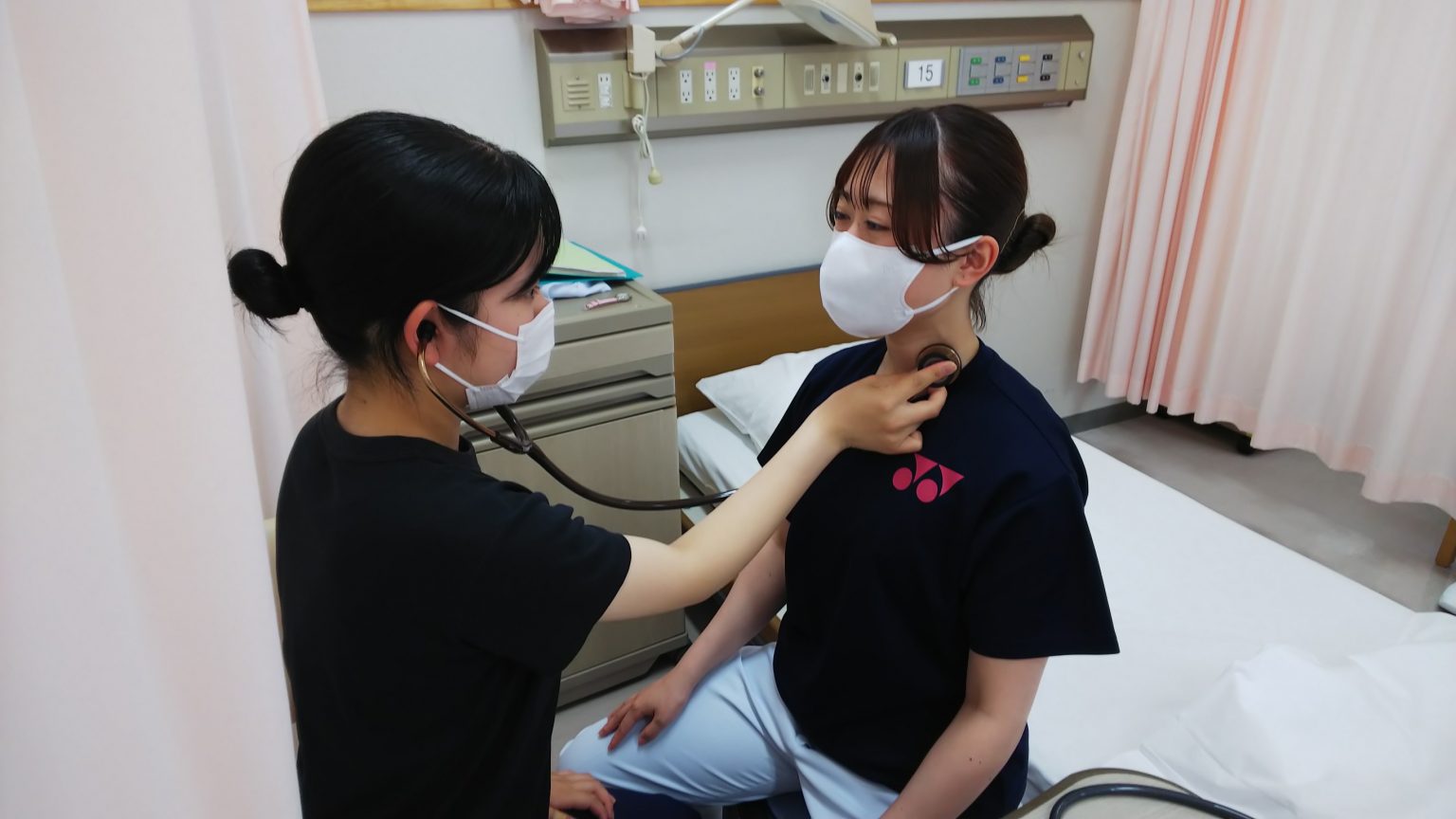 看護学生が患者に聴診器を使って心音を確認している様子。病室の中での実習風景。