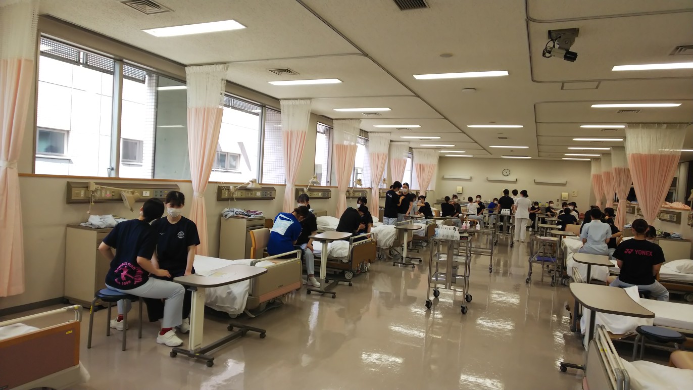 看護技術の実習を行う学生たちが並ぶ教室の様子。ベッドや医療器具が配置されている。