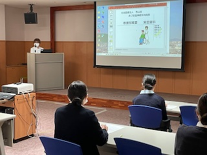 基礎看護学実習のオリエンテーションで、学生が講義を受けている様子。講師がスライドを使って説明している。