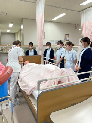 看護技術の演習で洗髪を学ぶ1年生の学生たちが、ベッドにいる人形を囲んでいる様子。