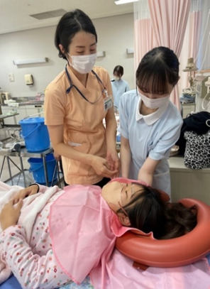 看護技術の演習で洗髪を行う学生たちが、患者役の女性に対してケアを施している様子。