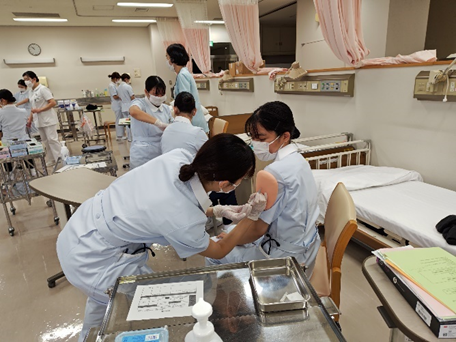 看護技術の演習で筋肉内注射を行う学生たちが、実習室で協力している様子。