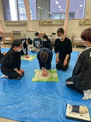看護技術の演習で心肺蘇生法を学ぶ学生たちが、青いシートの上で実習を行っている様子。