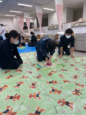 看護技術の演習で、学生たちが救命救急処置を学ぶ様子。床にはカラフルな模様のシートが敷かれている。