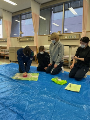 看護技術の演習で救命救急処置を学ぶ学生たちが、青いシートの上で実習を行っている様子。