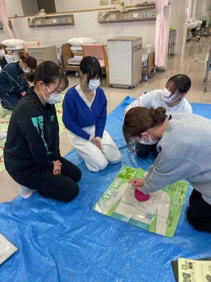 看護技術の演習で救命救急処置を学ぶ学生たちが、青いシートの上で実習を行っている様子。