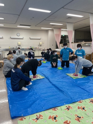 看護技術の演習で救命救急処置を学ぶ2年生の学生たちが、青いシートの上で真剣に取り組んでいる様子。