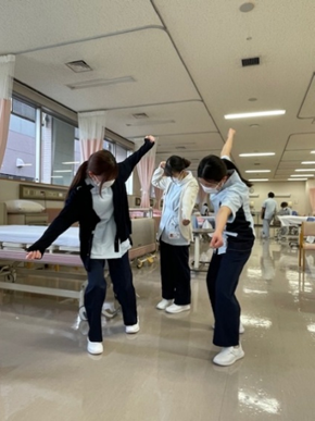 看護技術の自己学習を行う学生たちが、病院の実習室で楽しそうにポーズをとっている様子。