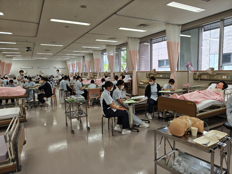 看護技術の演習室で、学生たちが経鼻経管栄養の管理を実践している様子。モデル人形がベッドに横たわっている。