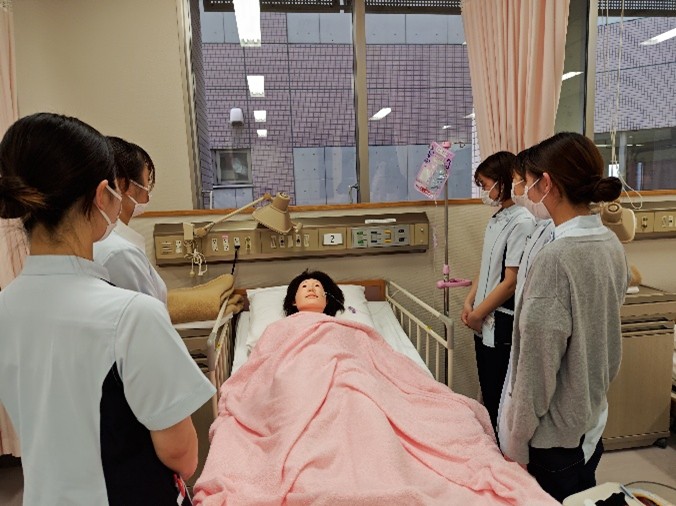 看護技術の演習で経管栄養を学ぶ学生たちが、ベッドに横たわる人形を囲んでいる様子。