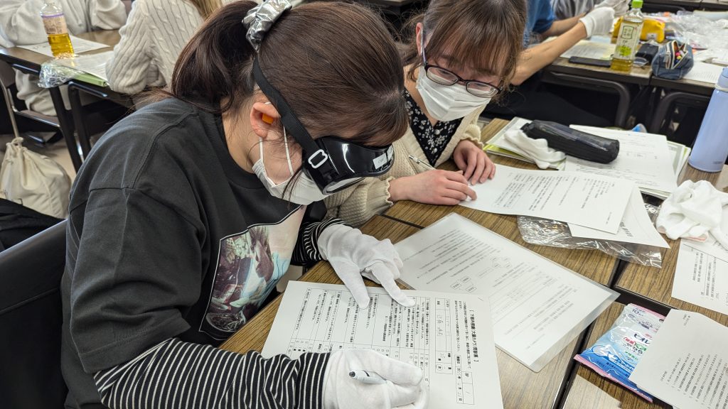 高齢者の疑似体験を行う学生たちが、食事介助やとろみ食体験に関する資料に取り組んでいる様子。