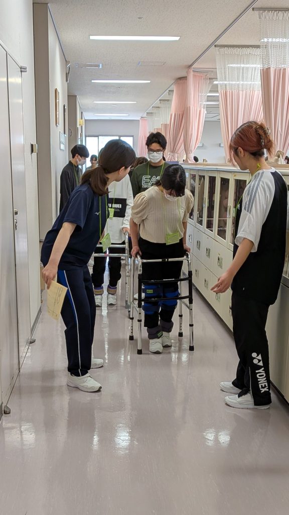 高齢者の疑似体験を行う学生たちが、歩行器を使って歩く様子を支援している場面。
