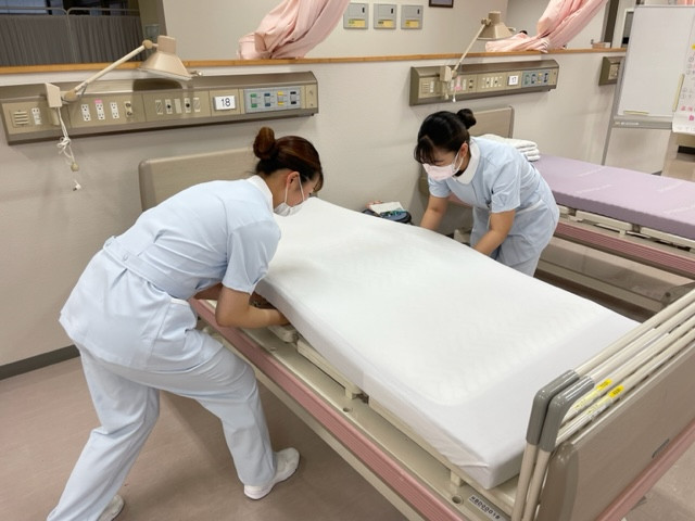看護学部の学生が病院のベッドにシーツを張っている様子。清潔感あふれる医療現場の一コマ。